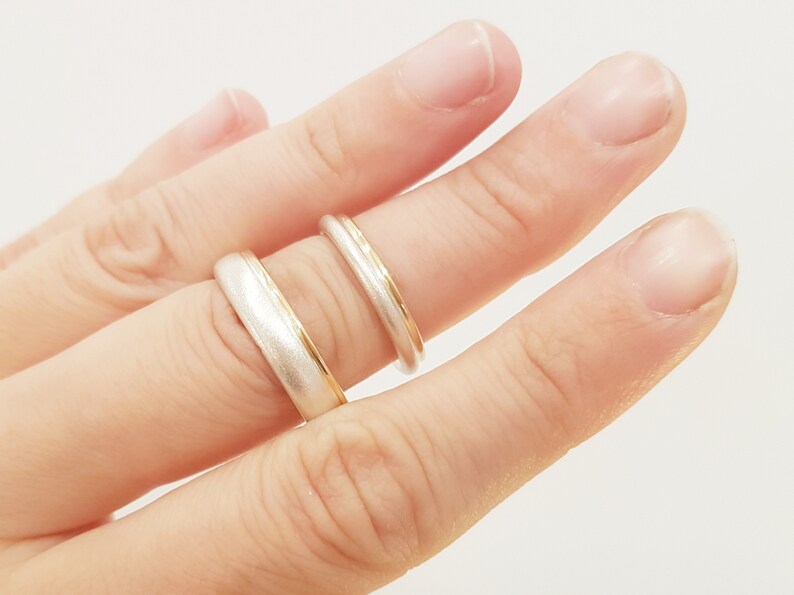 May include: Two wedding bands on a finger. The top band is a thin gold band with a white metal edge. The bottom band is a wider white metal band with a thin gold edge.