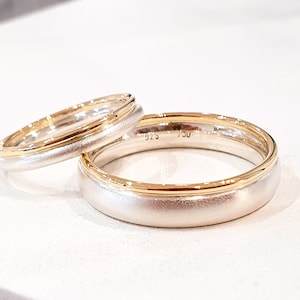 May include: Two wedding bands, one with a brushed silver finish and a thin gold band, the other with a brushed silver finish and a thicker gold band. The bands are inscribed with "925" and "750".