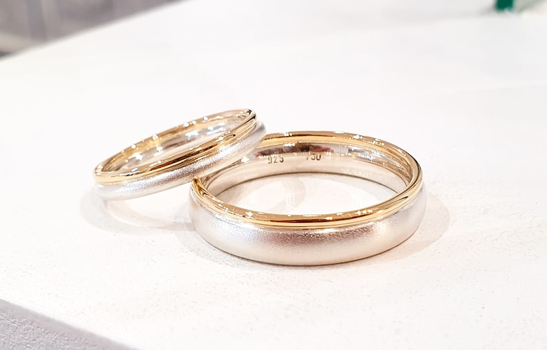 May include: Two wedding bands, one with a brushed silver finish and a thin gold band, the other with a brushed silver finish and a thicker gold band. The bands are inscribed with "925" and "750".