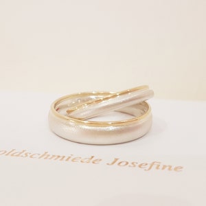 May include: Two wedding bands, one with a brushed white finish and a thin gold band, and the other with a brushed white finish and a gold band around the edge. The bands are stacked on top of each other.