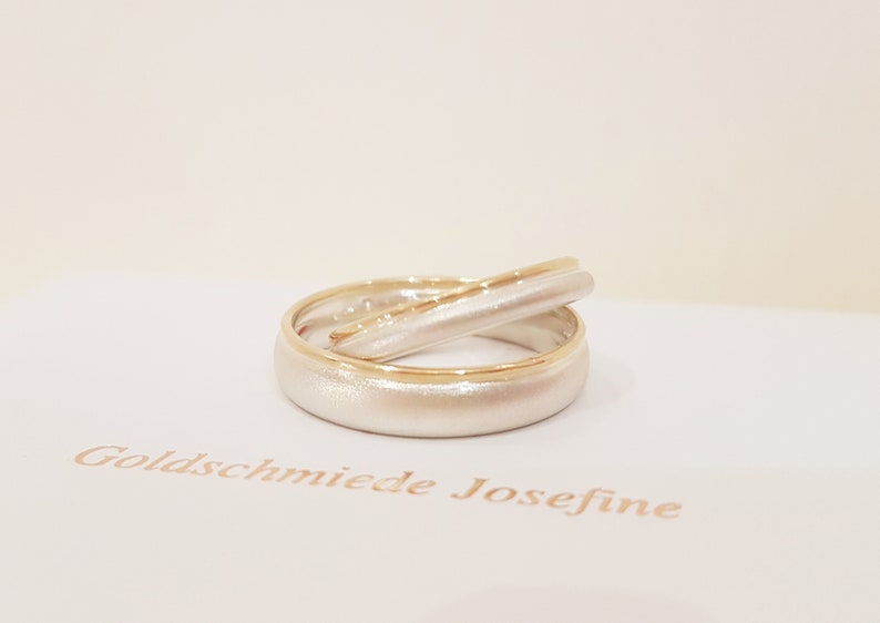 May include: Two wedding bands, one with a brushed white finish and a thin gold band, and the other with a brushed white finish and a gold band around the edge. The bands are stacked on top of each other.
