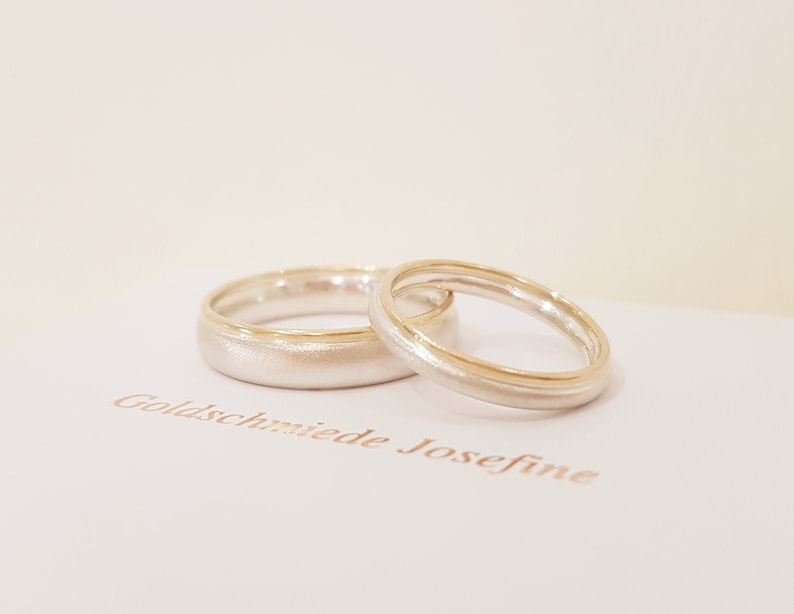 May include: Two wedding bands, one silver with a thin gold band around the edge, and the other silver with a thicker gold band around the edge. The bands are on a white surface with the text "Goldschmiede Josefine" printed in brown.