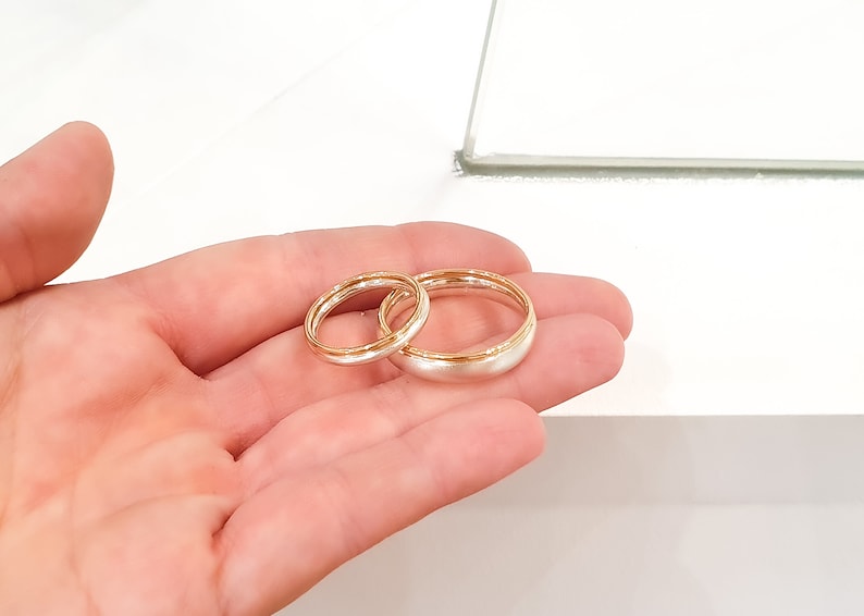 May include: Two wedding bands, one silver and one gold, are displayed on a hand. The bands are both simple and elegant.