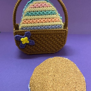 May include: A brown crocheted basket with a white ribbon handle and a multi-colored crocheted egg inside. The basket has a purple flower with a yellow center. The egg is sitting on a brown cork coaster with a blue crocheted edge.
