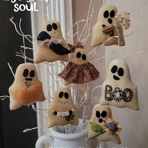 以下が含まれることがあります： 白い枝から吊るされた、黒い目と笑顔の口を持つ7つのベージュ色の布製ゴーストオーナメント。 各ゴーストには、カボチャ、カラス、「BOO」の文字など、ユニークなアクセサリーが付いています。 「a joyful soul」と「7 Little Ghosties Pattern」の文字が見えます。