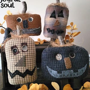 May include: Four handmade fabric pumpkins with stitched faces and button eyes. The pumpkins are in shades of brown, tan, and blue, with twig stems and straw accents. The text "a joyful soul" is in the upper left corner.