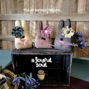 Peut inclure: Trois poupées lapins faites à la main, ornées de fleurs, sont posées sur une boîte en bois rustique. Les lapins sont beiges, bruns et gris, avec des visages cousus et des moustaches. La boîte porte l'inscription "a joyful soul".
