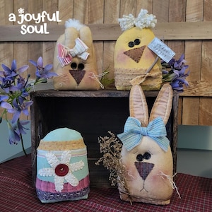 May include: Handmade Easter decorations including a bunny, chick, and egg. The bunny has a blue bow and the chick holds a sign that says "Happy Spring". The egg is decorated with a flower and button. The decorations are on a wooden crate.