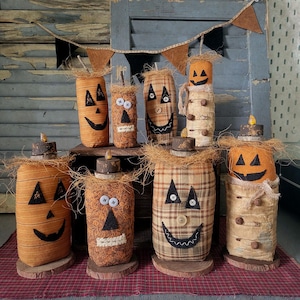 May include: A collection of eight fabric-covered cylindrical figures resembling jack-o'-lanterns, each featuring stitched felt faces, button eyes, and straw-like accents. The figures are arranged on wooden bases, with some topped by small wooden discs.