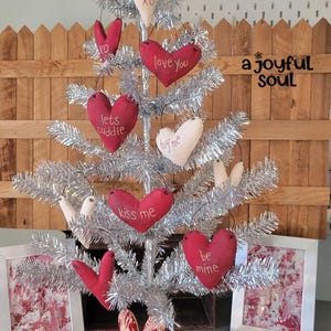 May include: A silver tinsel Christmas tree decorated with red and cream-colored heart ornaments. The hearts have phrases like "love you" and "kiss me" embroidered on them. Two framed pictures and three heart-shaped ornaments are at the base of the tree. The text "a joyful soul" is visible.