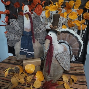 Op de afbeelding: Twee pluche kalkoenversieringen. Eén kalkoen draagt een blauwe sjaal met witte zigzags en een vleugel met tweedpatroon. De andere kalkoen draagt een bordeauxrode sjaal en een bruine hoed. Herfstbladeren en een houten blok staan op de voorgrond.