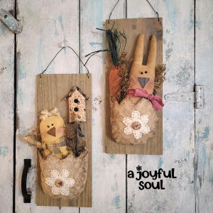 May include: Two rustic wooden wall hangings. One features a yellow chick in an egg-shaped pocket with a flower, and a small birdhouse. The other has a bunny with a carrot and a pink bow. Both are spring-themed.