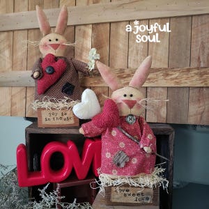 May include: Two fabric bunny ornaments, one in brown, the other in pink, holding hearts and flowers. The text "joy is in the air" and "love sweet love" are on small wooden signs. The background includes the text "a joyful soul".