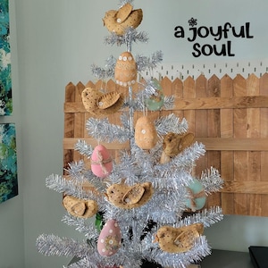 May include: A silver tinsel Christmas tree decorated with handmade ornaments. The ornaments include bird and egg shapes in various colors and patterns. The background features the text "a joyful soul" and a wooden fence.