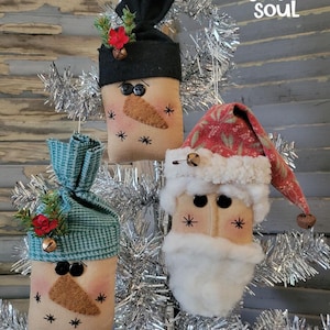 May include: Three fabric ornaments featuring two snowmen with black button eyes, stitched carrot noses, and hats in black and teal, and one Santa figure with a red patterned hat, white faux fur trim, and a white beard. Text reads a joyful soul.
