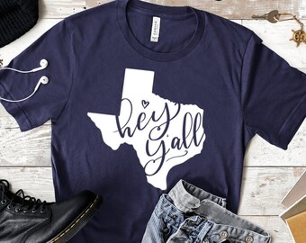 May include: A navy blue t-shirt with a white outline of the state of Texas and the text "hey yall" inside the outline. A small white heart is above the word "hey".