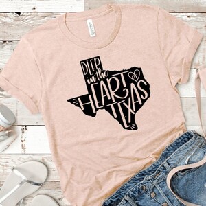 May include: A light pink t-shirt with a black graphic of the state of Texas. The graphic says "Deep in the Heart of Texas" with a heart symbol.