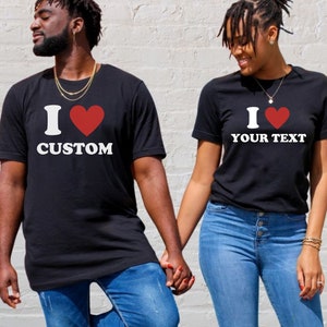 Puede incluir: Una pareja lleva camisetas negras con un coraz&oacute;n rojo y el texto "I love CUSTOM" y "I love YOUR TEXT". Se toman de la mano y sonr&iacute;en.