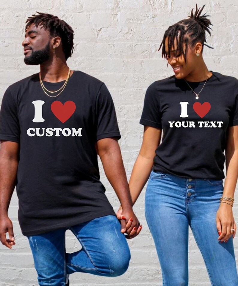 Puede incluir: Una pareja lleva camisetas negras con un coraz&oacute;n rojo y el texto "I love CUSTOM" y "I love YOUR TEXT". Se toman de la mano y sonr&iacute;en.