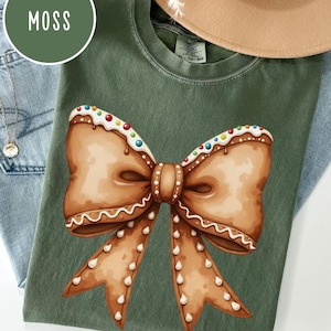 May include: A green t-shirt with a gingerbread bow design. The bow is decorated with white icing and colorful sprinkles. The shirt has the text "Simple Colors" and "XL - TO - EG" printed on the tag.