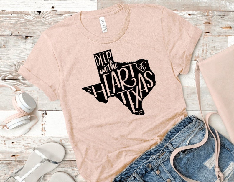 May include: A light pink t-shirt with a black graphic of the state of Texas. The graphic says "Deep in the Heart of Texas" with a heart symbol.