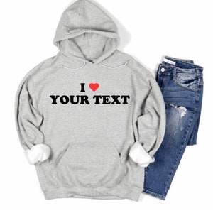 Puede incluir: Sudadera gris con capucha con un gr&aacute;fico negro que dice "I love YOUR TEXT" con un coraz&oacute;n rojo.