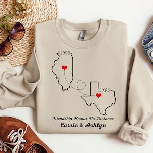 May include: Tan sweatshirt with a black outline of Illinois and Texas with red hearts on each state. The text "Friendship Knows No Distance Carrie & Ashlyn" is printed below the states.