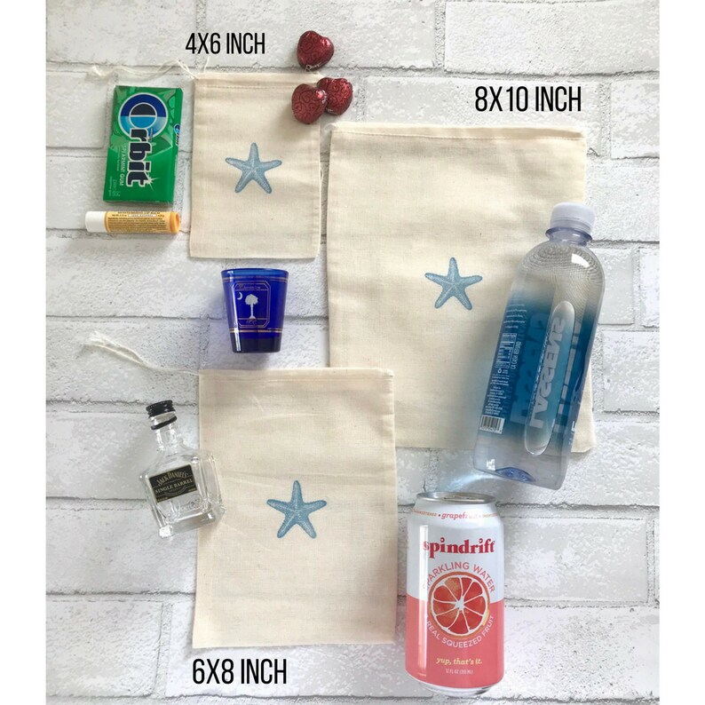 May include: Four different sizes of natural cotton muslin bags with a blue starfish design. The bags are 4x6 inches, 6x8 inches, and 8x10 inches.  The bags are displayed on a white brick wall with other items, including a bottle of water, a can of sparkling water, a small bottle of liquor, a pack of gum, a tube of lip balm, and a small blue shot glass.