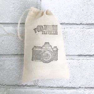May include: A white cotton drawstring bag with a black and white image of a camera and the text "World Traveler" printed on it.
