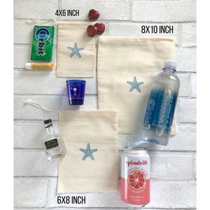 May include: Four different sizes of natural cotton muslin bags with a blue starfish design. The bags are 10x15 cm, 15x20 cm, and 20x25 cm. The bags are displayed on a white brick wall with other items, including a bottle of water, a can of sparkling water, a small bottle of liquor, a pack of gum, a tube of lip balm, and a small blue shot glass.