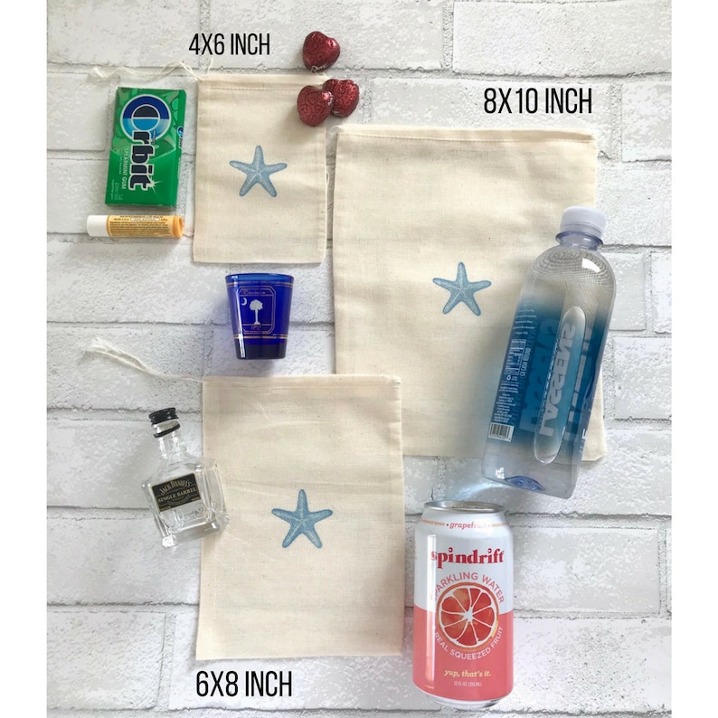 May include: Four different sizes of natural cotton muslin bags with a blue starfish design. The bags are 10x15 cm, 15x20 cm, and 20x25 cm. The bags are displayed on a white brick wall with other items, including a bottle of water, a can of sparkling water, a small bottle of liquor, a pack of gum, a tube of lip balm, and a small blue shot glass.