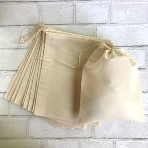 May include: A stack of 15 natural cotton muslin bags with drawstring closures. The bags are folded and stacked on a white brick background.