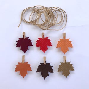May include: Six autumn-themed maple leaf clothes pegs in various colours, including red, orange, brown, and tan. The clothes pegs are attached to a length of twine.