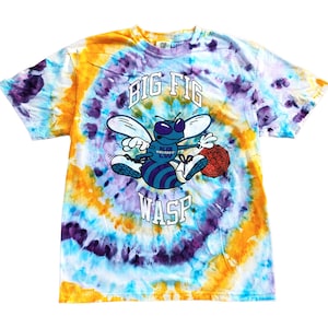 May include: A short-sleeve t-shirt with a spiral tie-dye pattern in yellow, purple, and light blue. The center features a cartoon wasp graphic with the text BIG FIG WASP and KG WOOOOO LW on the wasp's body, along with a red geometric sphere.