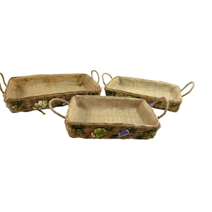 May include: Three rectangular woven baskets with handles. The baskets are made of natural-colored woven material and are decorated with floral and leaf accents. The handles are made of twisted rope.