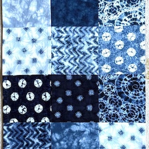 May include: A rectangular, indigo blue patchwork textile with a variety of patterns. The patterns include polka dots, spirals, and geometric designs. The textile is likely a table runner or decorative piece.