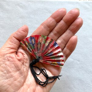 May include: A small red, gold, and blue paper fan with a black tassel. The fan is folded and has a floral pattern.
