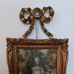 Può includere: Una stampa incorniciata con una cornice dorata ornata e un fiocco dorato decorativo nella parte superiore. La stampa raffigura una scena con figure in abiti d'epoca in un ambiente di giardino. La cornice è larga circa 15 cm.