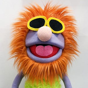 May include: A handmade puppet with a purple face, arms, and feet, and a bright orange furry head. The puppet has black sunglasses, a pink nose, and a green furry chest. The puppet is standing on a white stand.