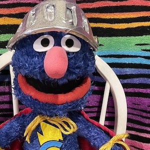 May include: Blue furry puppet wearing a red cape, a yellow lightning bolt with a blue "G" on its chest, and a silver helmet. The puppet is sitting on a white chair.