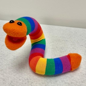 May include: A plush toy shaped like a worm with a bright orange head and two black plastic eyes. The body is covered in horizontal stripes of rainbow colors including red, yellow, green, blue, and purple. The mouth is open, revealing a pink interior.