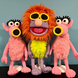 May include: Three colorful plush puppets. The central puppet has orange fur, yellow sunglasses, and a purple face. Two pink puppets with large eyes and yellow-rimmed mouths flank the central puppet. All puppets have a fuzzy texture and are standing on white bases.