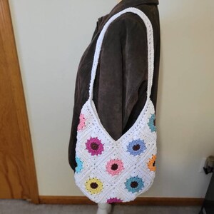 May include: White crocheted shoulder bag with a colorful flower pattern. The bag has a long strap and is made of white yarn with colorful flowers crocheted into the design.