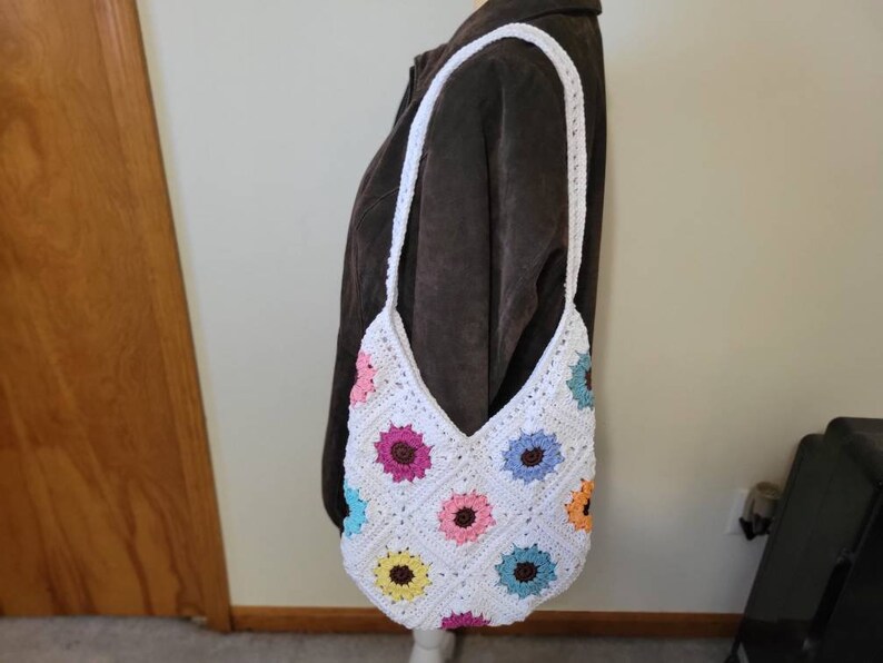 May include: White crocheted shoulder bag with a colorful flower pattern. The bag has a long strap and is made of white yarn with colorful flowers crocheted into the design.