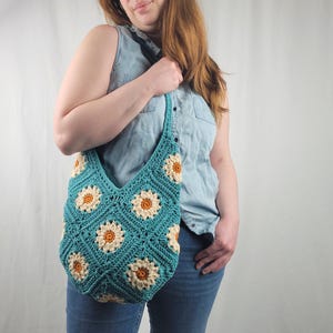 May include: A teal crocheted tote bag with a daisy pattern. The bag has a long strap and is worn over the shoulder.