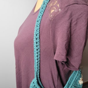 May include: A teal blue crocheted shoulder bag with a long strap. The bag is being worn by a person wearing a maroon shirt.