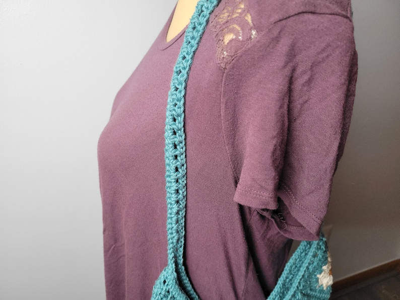 May include: A teal blue crocheted shoulder bag with a long strap. The bag is being worn by a person wearing a maroon shirt.