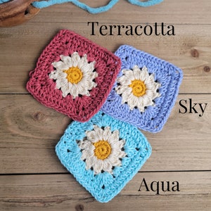 May include: Three crocheted squares with a daisy design in the center. The squares are in terracotta, sky blue, and aqua colors. The text "Terracotta", "Sky", and "Aqua" are written below each square.