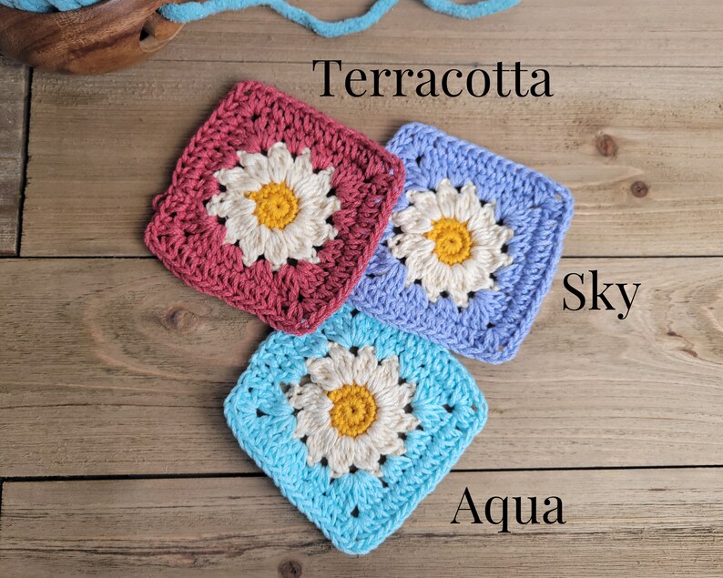May include: Three crocheted squares with a daisy design in the center. The squares are in terracotta, sky blue, and aqua colors. The text "Terracotta", "Sky", and "Aqua" are written below each square.