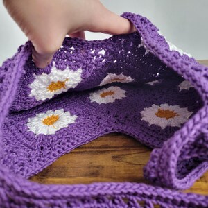May include: Purple crochet tote bag with white daisy flower accents. The bag is open and the inside is visible.